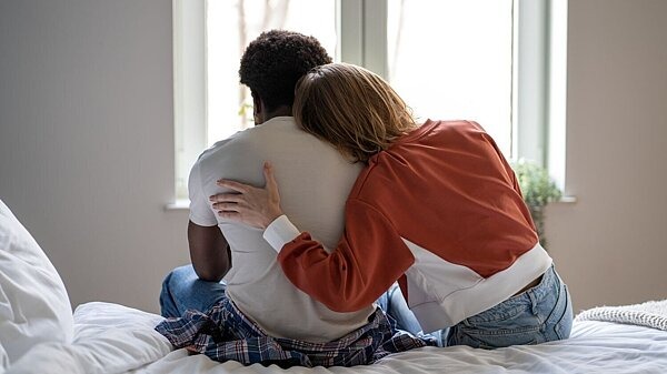 Eine Person legt den Arm um ihren Partner, während beide nebeneinander auf einem Bett sitzen. Symbolbild für emotionale Belastung und Unterstützung in Beziehungen bei Glücksspielsucht.