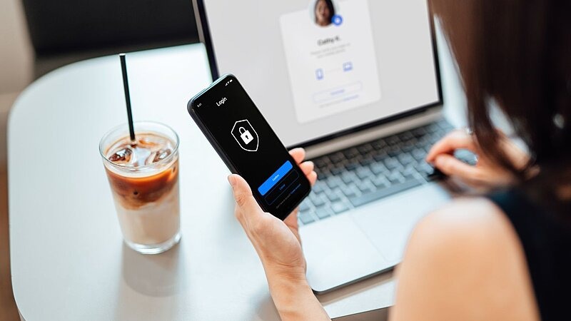 Eine Person hält ein Smartphone mit einer Sicherheitsfunktion in der Hand, während sie an einem Laptop sitzt – symbolisch für schützen eigener Finanzen., Bild: Oscar Wong / gettyimages